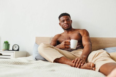 Photo for Confident muscular african american man in pajama pants with coffee cup looking at camera in bedroom - Royalty Free Image