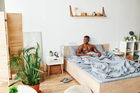 Photo for Shirtless muscular african american man sitting on bed in spacious bedroom with green potted plants - Royalty Free Image