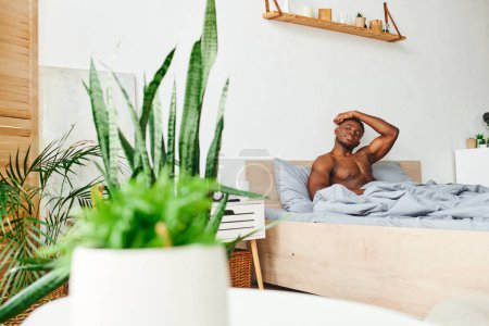 Photo for Muscular handsome african american man sitting on bed in spacious bedroom with green potted plants - Royalty Free Image