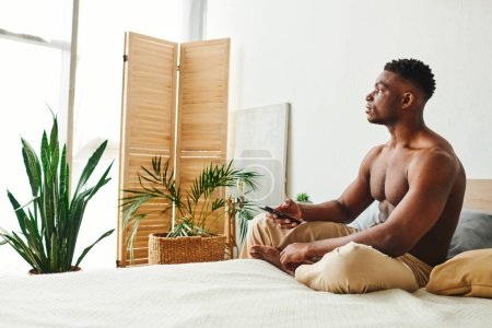 dreamy african american man in pajama pants sitting on bed with smartphone and looking away