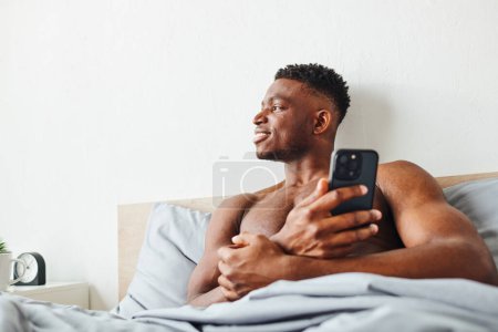 cheerful muscular african american with mobile phone looking away in modern bedroom on cozy bed
