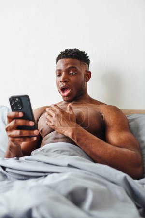 amazed muscular african american man looking at smartphone while browsing internet in bedroom