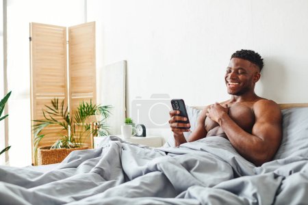 laughing shirtless african american man looking at smartphone while browsing internet in bedroom