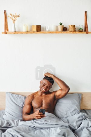 cheerful athletic african american man browsing social medial on smartphone and laughing in bedroom