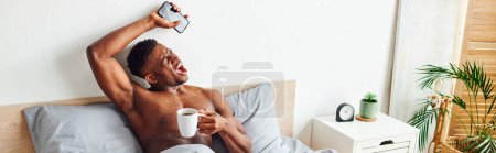muscular african american man with coffee cup and smartphone yawning and stretching on bed, banner