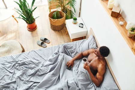 top view of muscular african american guy with coffee cup and smartphone looking away on bed