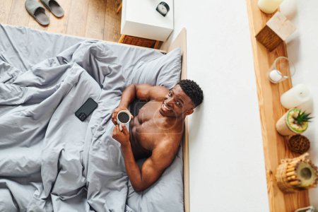 top view of cheerful shirtless african american man with morning coffee smiling at camera on bed