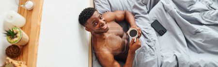 top view of happy muscular african american man with morning coffee looking at camera on bed, banner