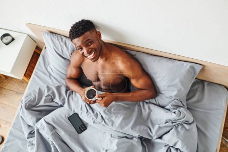 top view of carefree athletic african american man with morning coffee looking at camera on bed