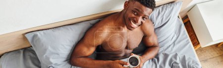top view of young joyous african american man with coffee cup smiling at camera on bed, banner