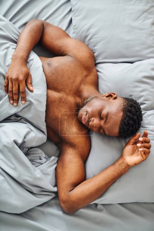 Téléchargez les photos : Vue du dessus de jeune homme afro-américain gars dormir sur un lit confortable sous une couverture douce le matin - en image libre de droit