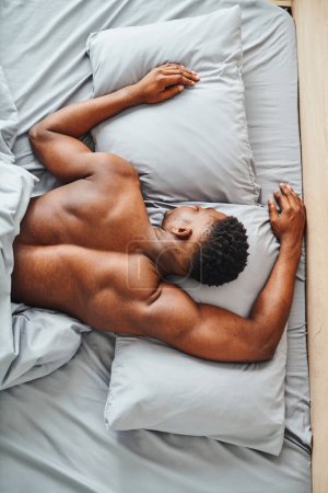 Téléchargez les photos : Vue de dessus du jeune homme afro-américain dormant sur la literie grise dans la chambre moderne, loisirs matinaux - en image libre de droit