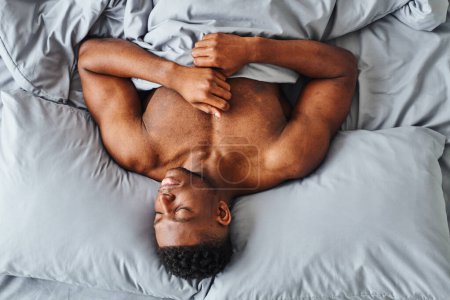 Téléchargez les photos : Vue de dessus de l'homme afro-américain avec le corps fort rêvant sur la literie confortable grise à la maison - en image libre de droit