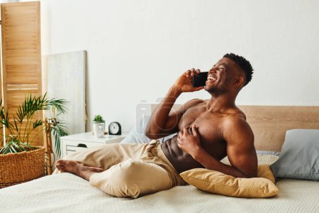 cheerful shirtless african american man in pajama pants talking on smartphone in modern bedroom
