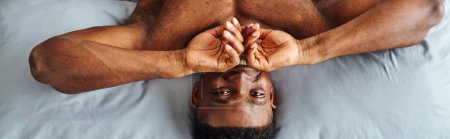 Photo for Joyful african american guy covering mouth and smiling at camera waking up on bed, top view, banner - Royalty Free Image