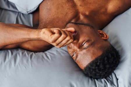 Foto de Vista superior del soñoliento hombre afroamericano bostezando y mirando hacia otro lado mientras se despierta por la mañana en casa - Imagen libre de derechos