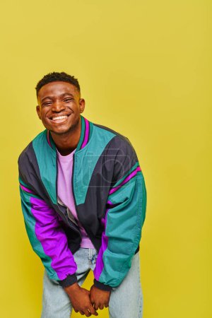überglücklich trendiger afrikanisch-amerikanischer Mann in heller Windjacke lächelt und posiert auf gelbem Hintergrund