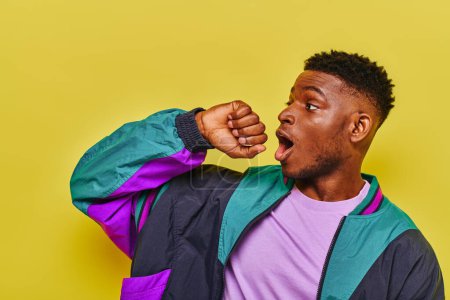 Téléchargez les photos : Homme afro-américain étonné avec bouche ouverte dans une veste coupe-vent à la mode regardant loin sur jaune - en image libre de droit