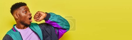 Téléchargez les photos : Homme afro-américain à la mode étonné dans une veste coupe-vent élégant regardant loin sur jaune, bannière - en image libre de droit