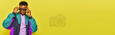 Téléchargez les photos : Confiant homme afro-américain en blouson coupe-vent lumineux ajuster les lunettes de soleil sur jaune, bannière - en image libre de droit