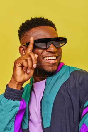 Téléchargez les photos : Homme afro-américain joyeux dans une veste coupe-vent ajuster les lunettes de soleil et détourner les yeux sur jaune - en image libre de droit