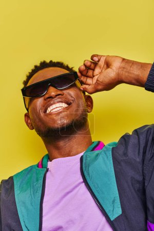 Téléchargez les photos : Joyeux afro-américain en coupe-vent veste lunettes de soleil posant avec un sourire radieux sur jaune - en image libre de droit