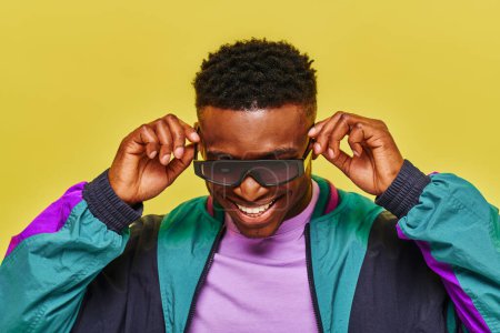 Téléchargez les photos : Homme africain élégant en veste colorée ajuster les lunettes de soleil et sourire sur fond jaune - en image libre de droit