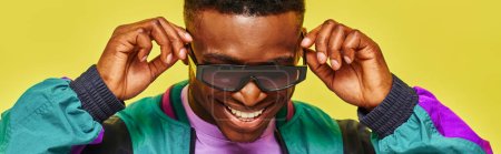 Téléchargez les photos : Homme africain à la mode en veste lumineuse ajuster les lunettes de soleil et sourire sur fond jaune, bannière - en image libre de droit