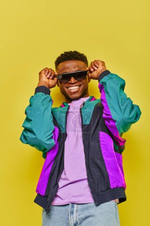 Téléchargez les photos : Homme afro-américain ravi en veste lumineuse et des lunettes de soleil à la mode souriant à la caméra sur jaune - en image libre de droit