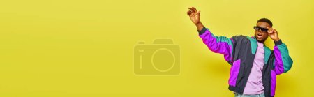 Téléchargez les photos : Homme afro-américain ravi en lunettes de soleil et veste lumineuse agitant la main sur fond jaune, bannière - en image libre de droit
