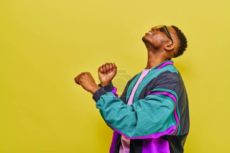 Téléchargez les photos : Jeune homme afro-américain branché en veste colorée et lunettes de soleil dansant sur jaune - en image libre de droit
