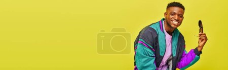 Téléchargez les photos : Homme afro-américain joyeux en veste lumineuse avec des lunettes de soleil regardant la caméra sur jaune, bannière - en image libre de droit