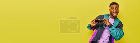Téléchargez les photos : Homme afro-américain souriant en veste lumineuse tenant des lunettes de soleil et dansant sur jaune, bannière - en image libre de droit