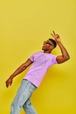 cheerful african american guy in sunglasses and purple t-shirt showing victory sign on yellow