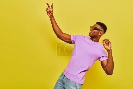 Téléchargez les photos : Homme afro-américain souriant dans des lunettes de soleil élégantes et t-shirt violet montrant signe de victoire sur jaune - en image libre de droit