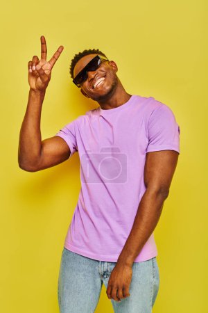 Téléchargez les photos : Heureux homme afro-américain en tenue d'été à la mode et des lunettes de soleil montrant signe de victoire sur jaune - en image libre de droit