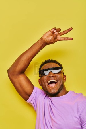 Téléchargez les photos : Ravi homme afro-américain élégant dans des lunettes de soleil montrant signe de victoire et crier sur jaune - en image libre de droit