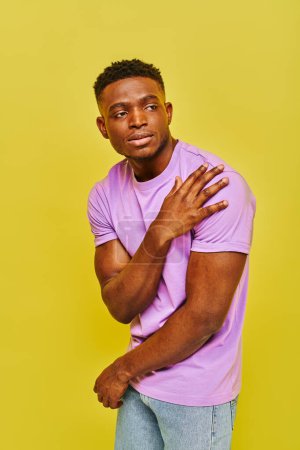 Téléchargez les photos : Jeune bel homme afro-américain en t-shirt violet posant et regardant loin sur fond jaune - en image libre de droit