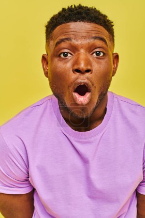 Téléchargez les photos : Homme afro-américain étonné en t-shirt violet avec bouche ouverte en regardant la caméra sur fond jaune - en image libre de droit