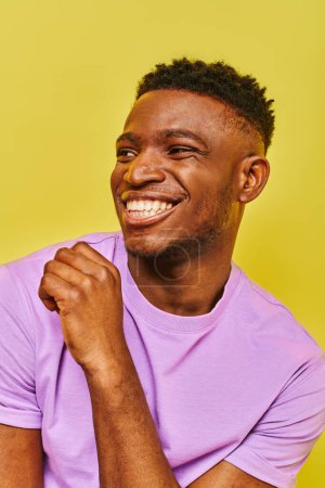 Foto de Despreocupado hombre afroamericano en camiseta púrpura sonriendo y mirando hacia otro lado en el fondo amarillo - Imagen libre de derechos
