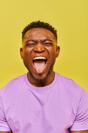 young african american man with closed eyes grimacing and sticking out tongue on yellow backdrop