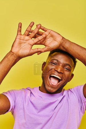 cheerful and funny african american man gesturing and sticking out tongue on yellow backdrop