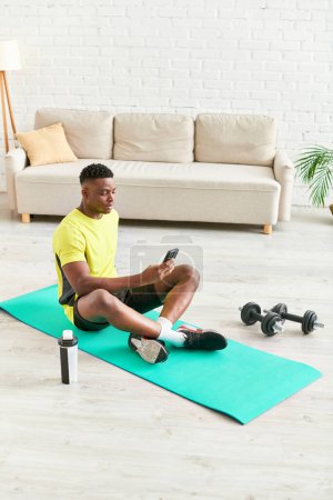 junger afrikanisch-amerikanischer Mann in Sportbekleidung sitzt mit Smartphone in der Nähe von Hanteln auf Fitnessmatte