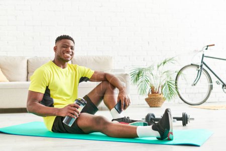 lächelnder Afroamerikaner in stylischer Sportbekleidung auf Fitnessmatte mit Sportflasche und Smartphone