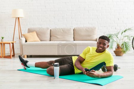 fröhlicher afrikanisch-amerikanischer Mann mit Smartphone blickt in Kamera auf Fitnessmatte im Wohnzimmer