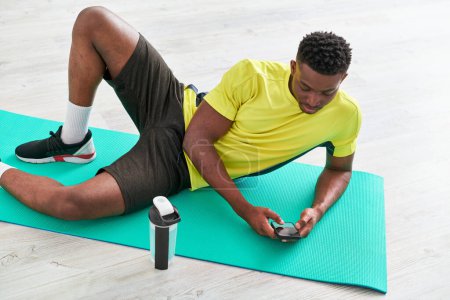 junge athletische afrikanisch-amerikanische Mann Nachrichten auf Smartphone auf Fitnessmatte in der Nähe von Sportflasche
