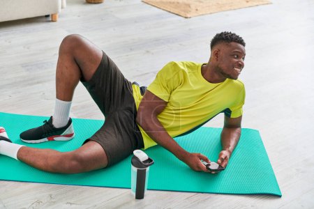 fröhlicher sportlicher afrikanisch-amerikanischer Mann mit Handy schaut auf Fitnessmatte in der Nähe von Sportflasche weg