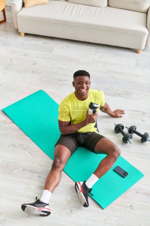 fröhlicher afrikanisch-amerikanischer Mann mit Sportflasche lächelt auf Fitnessmatte neben Hanteln in die Kamera