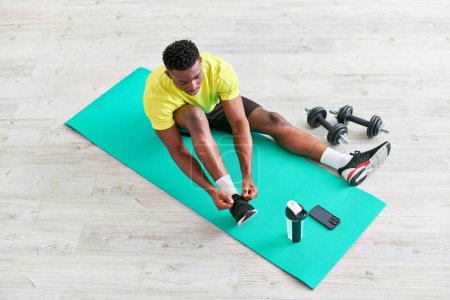 joven afroamericano hombre atando zapatilla de deporte en la alfombra de fitness cerca de teléfono inteligente y equipo deportivo