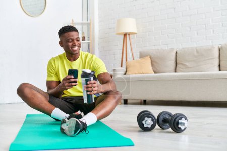 alegre hombre afroamericano con botella deportiva navegar por las redes sociales en el teléfono inteligente en la alfombra de fitness